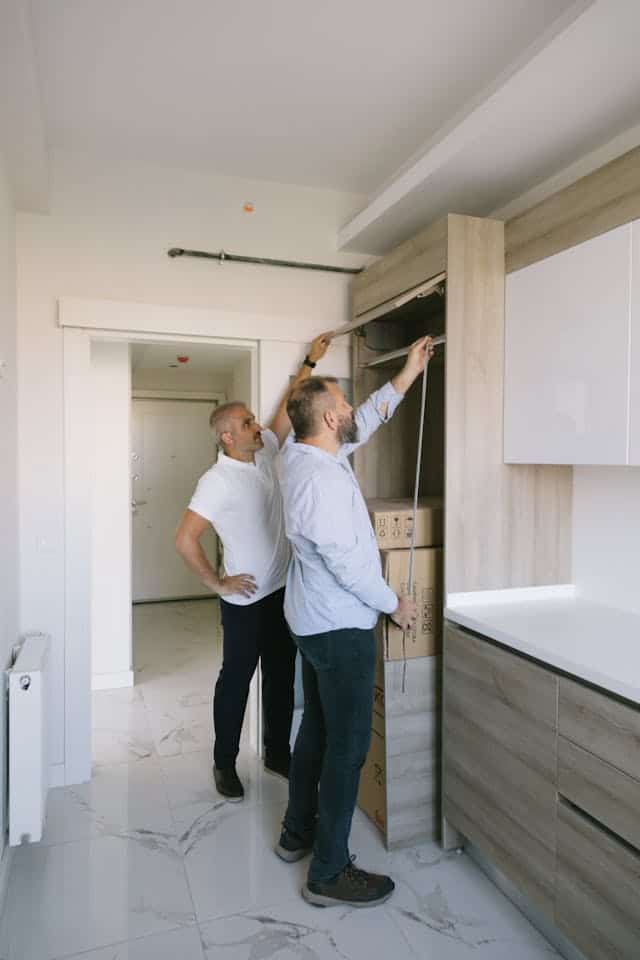 Two workers measuring cabinetry and inspecting interior fixtures, representing planning and assessment of building systems.