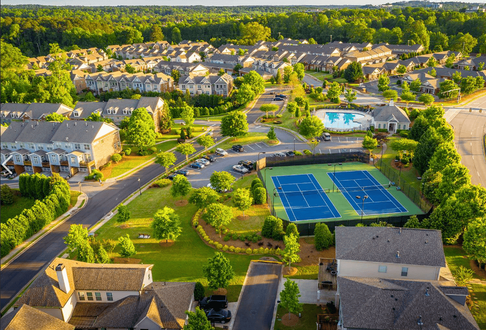 Aerial view of a planned residential community with rows of townhouses, tree-lined streets, parking areas, a fenced tennis court, and a shared swimming pool surrounded by greenery.