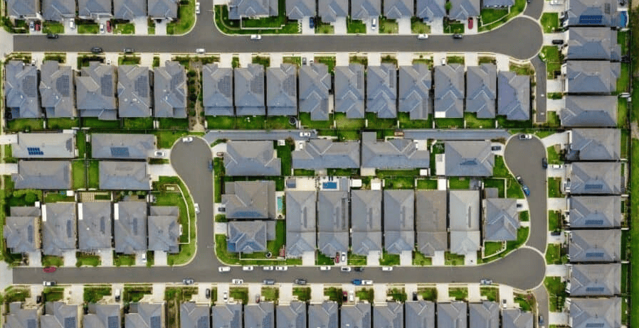 Top-down aerial view of a suburban housing development showing rows of closely spaced houses, curved residential streets, driveways, parked cars, and small green lawns arranged in a grid-like pattern.