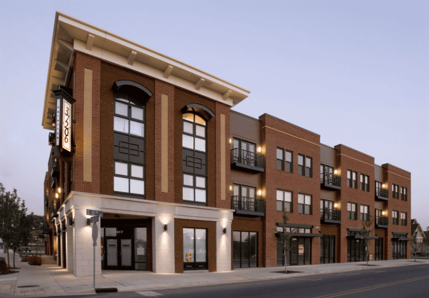 Street-level view of a modern mixed-use brick building with ground-floor retail spaces and upper-floor apartments featuring large windows and small balconies at dusk.